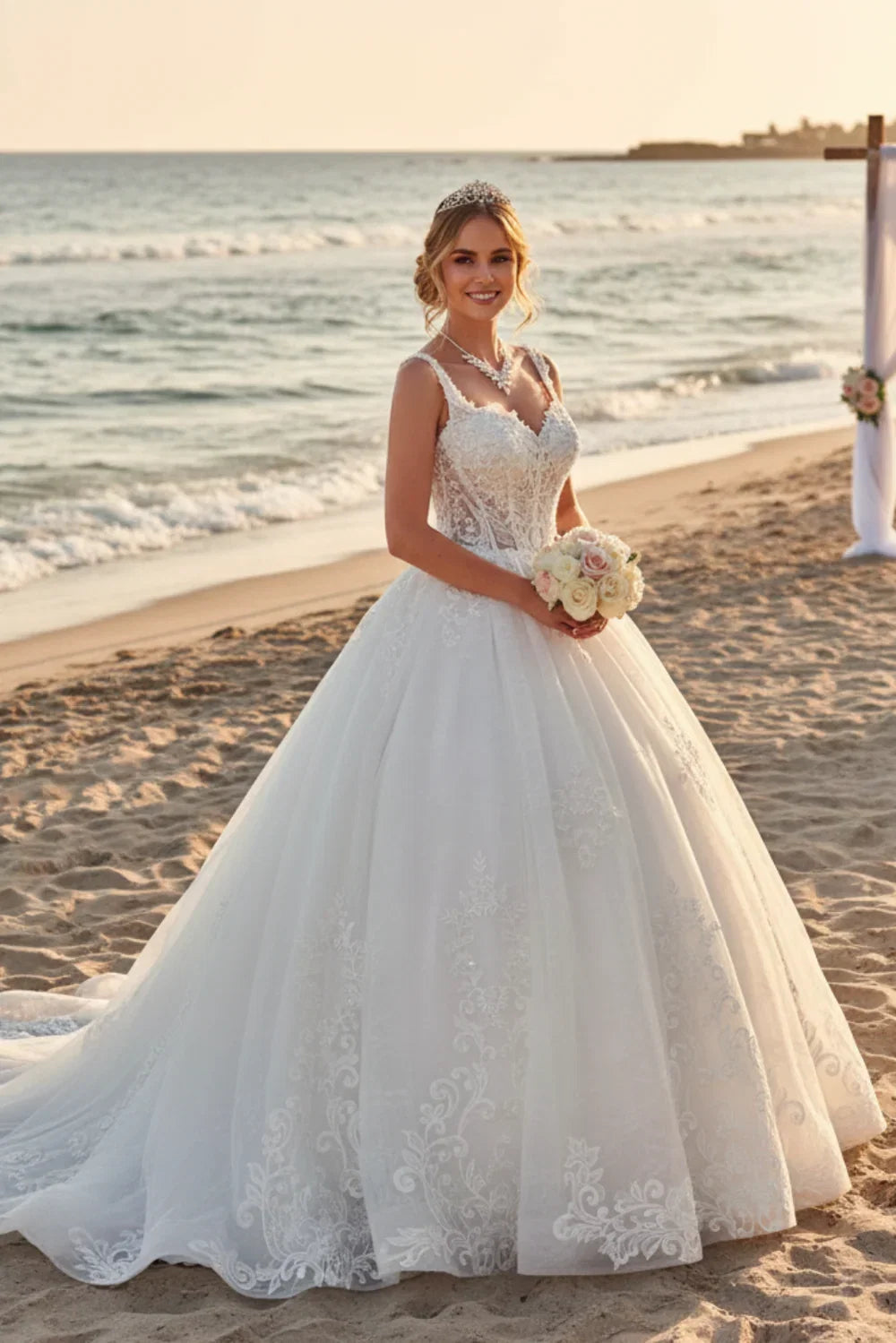A woman wearing a white lace wedding gown stands on a sandy beach holding a bouquet, with the ocean and sunset in the background.