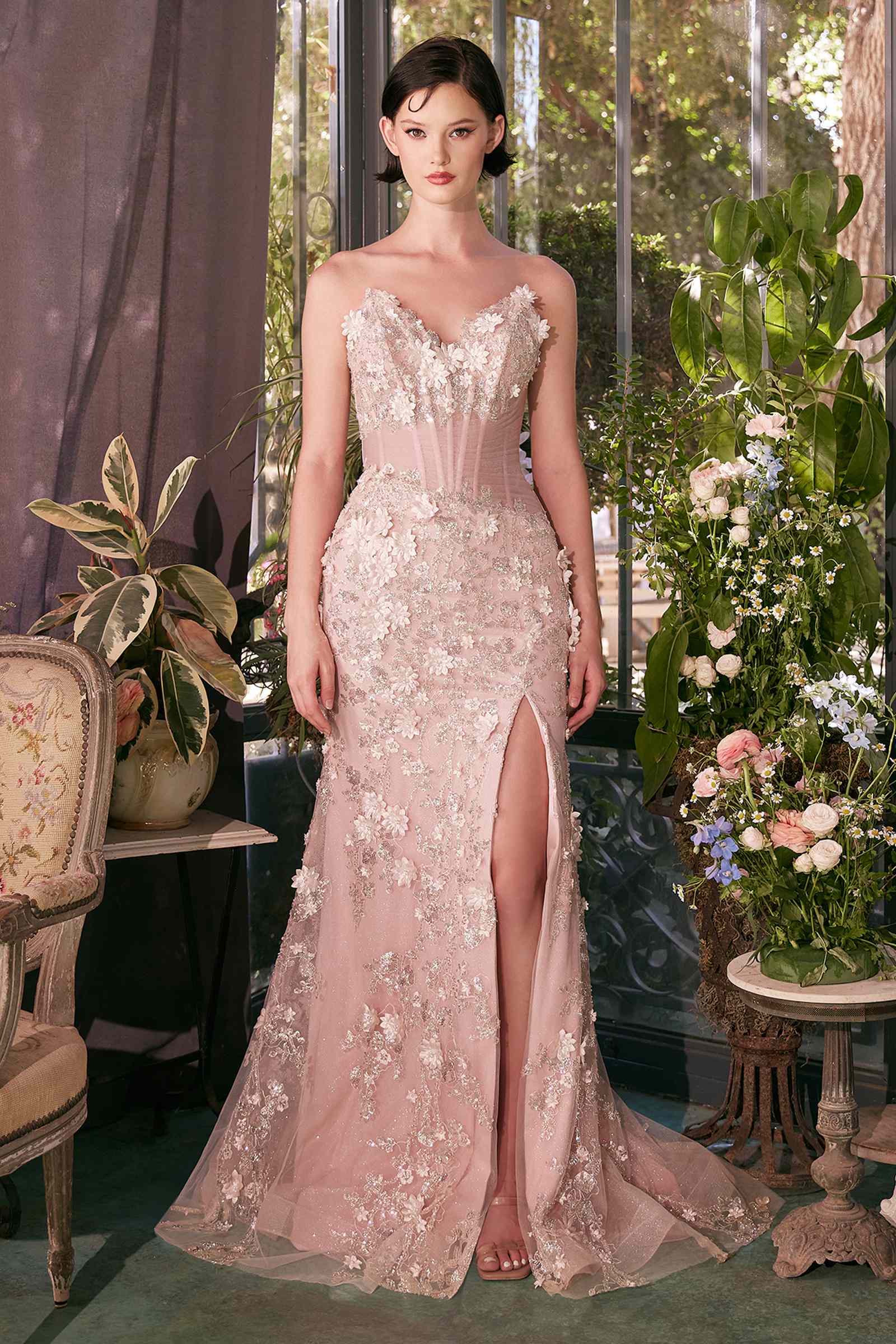 Woman in a pink floral evening gown standing in a decorated room with flowers and furniture.