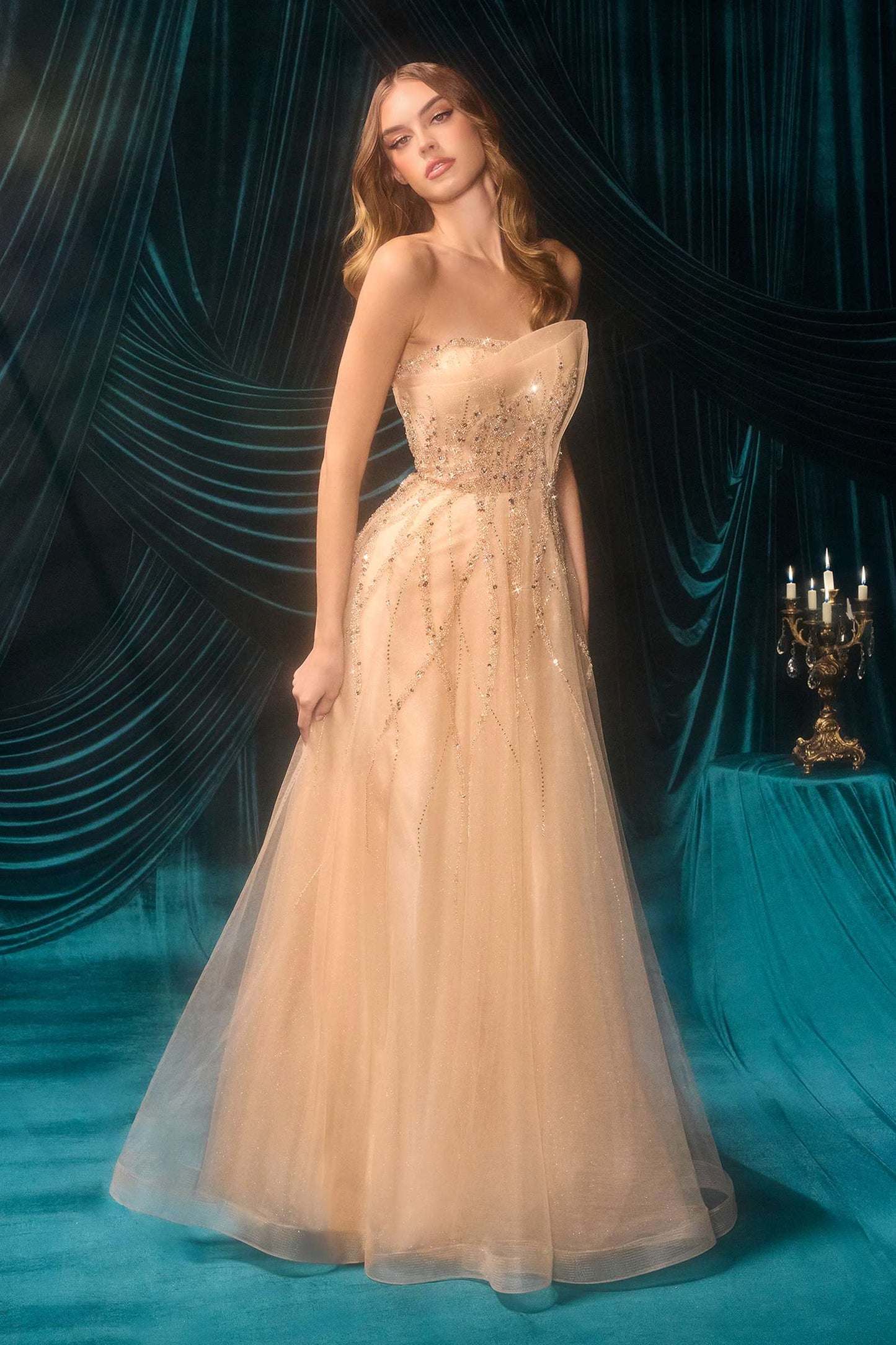Woman in a beige evening gown with intricate detailing against a dark curtain backdrop.