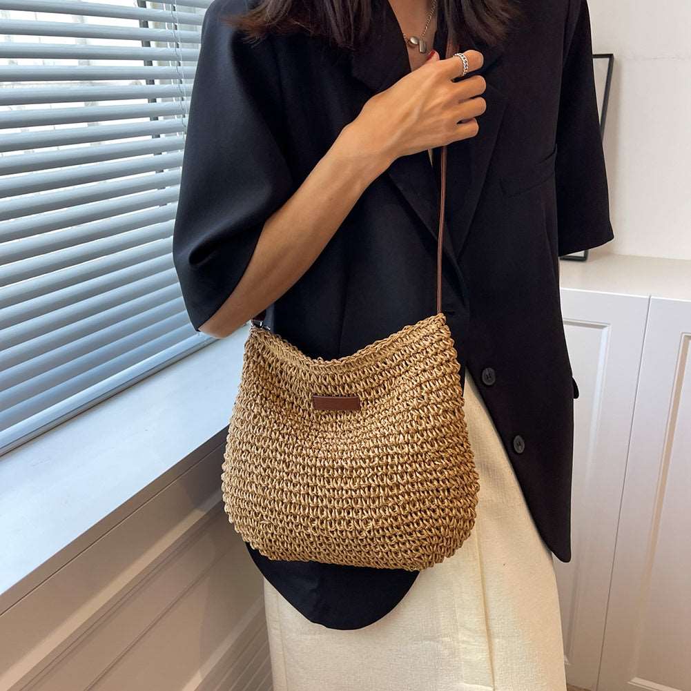 By a window with blinds, someone in a black blazer and white skirt holds the HalleBeauty Vacation Straw Tote Bag. This minimalistic, stylish outfit, accented by the bags leather patch and straw material, showcases modern fashion ideal for beach travel.