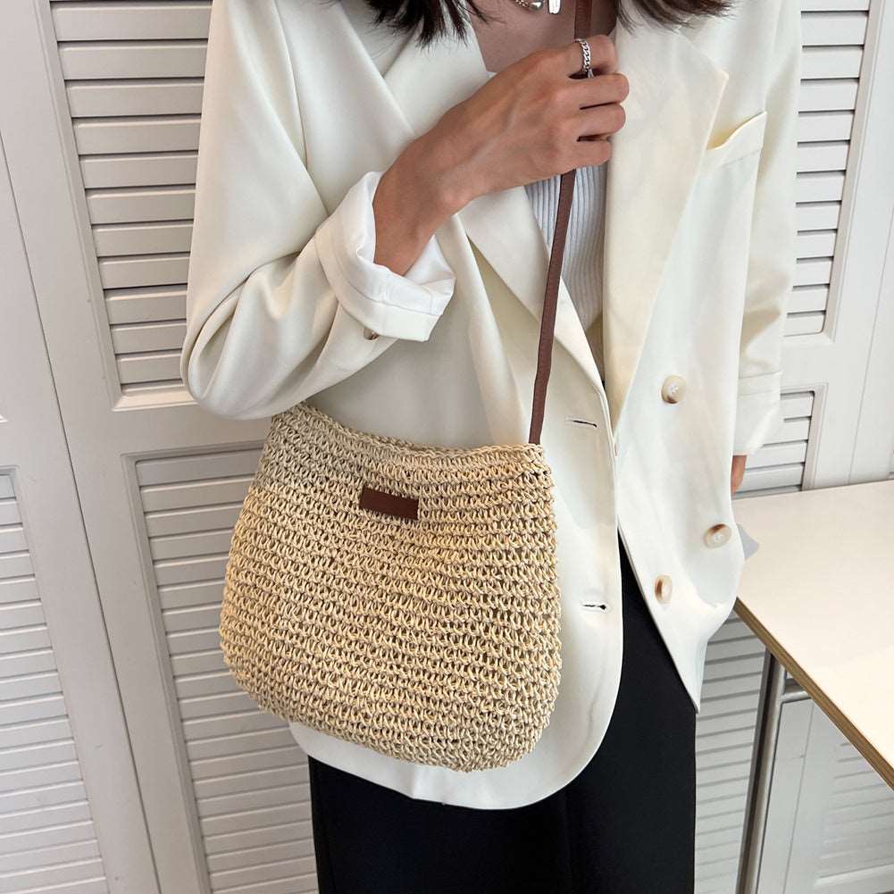 In front of white louvered doors, a person in a white blazer and black pants holds the HalleBeauty Vacation Straw Tote Bag | Stylish & Spacious Beach Bag for Travel, with its shoulder strap hinting at beach trips and sunny adventures.