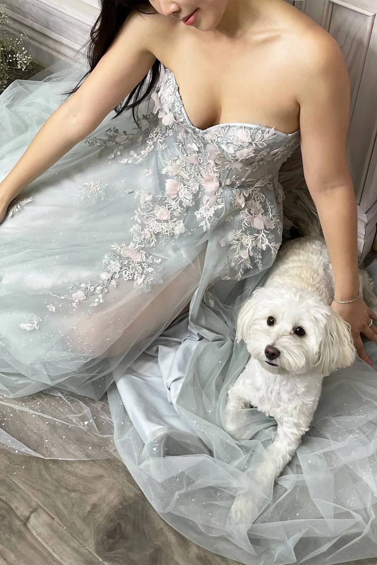 Woman in a light blue floral dress sitting with a small white dog on a wooden floor.