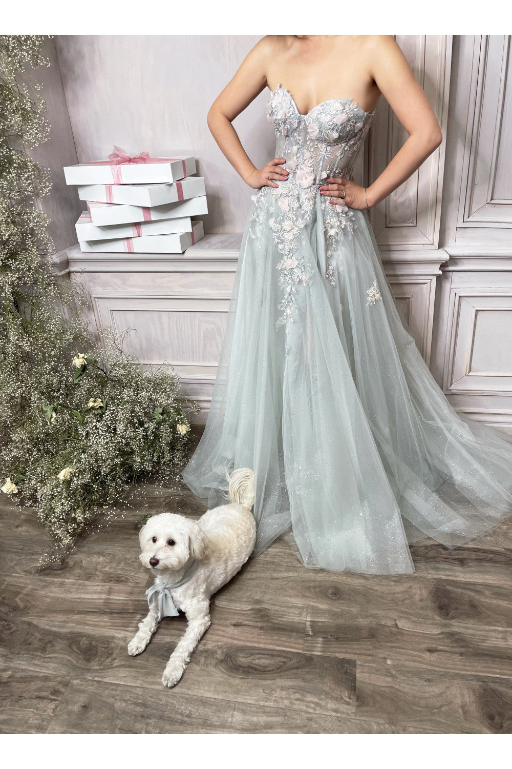 Woman in a light blue evening gown with floral details standing next to a white dog on a wooden floor.